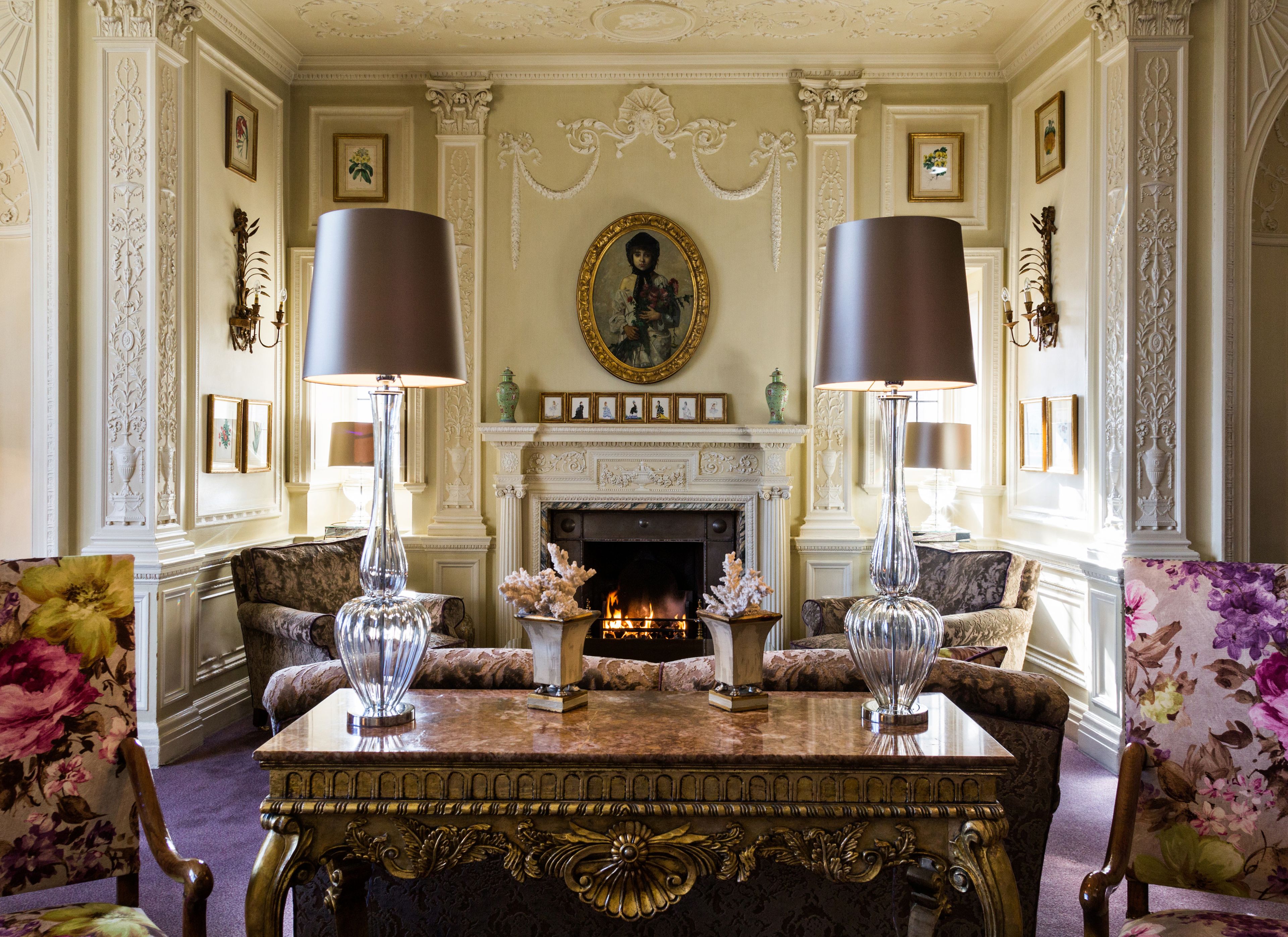 A luxurious room with a fireplace, two lamps, a table, and floral-patterned chairs.