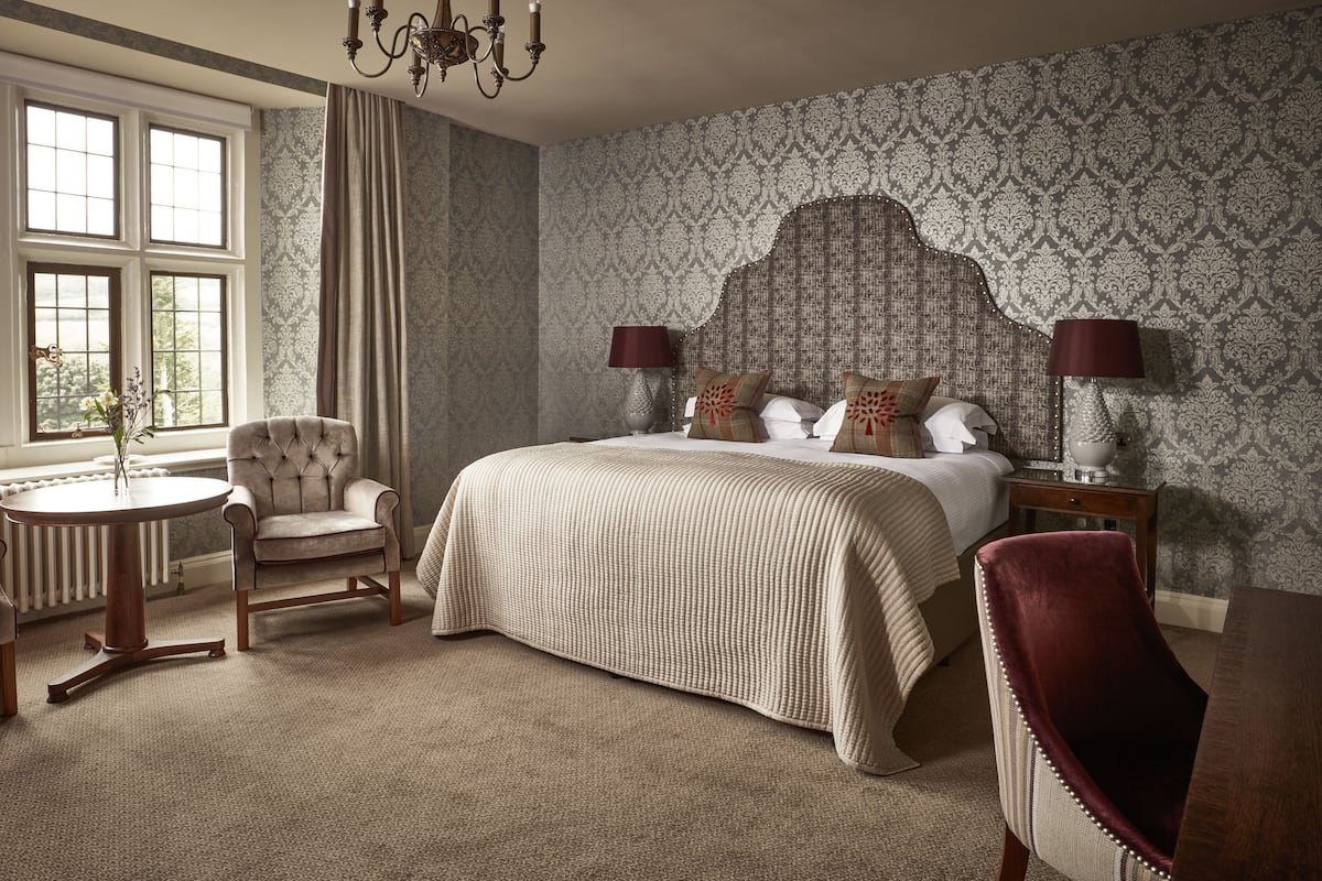 A bedroom with a large bed, a sitting area with a chair and a small table, and a view of greenery through the windows.