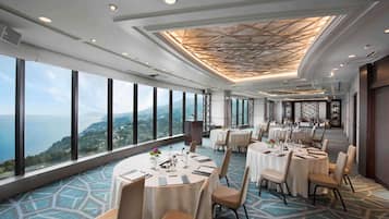 A high-ceilinged dining area with round tables, large windows offering a scenic view, and a patterned carpet.
