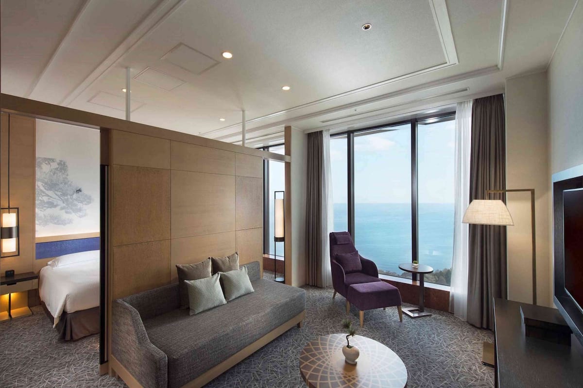 A modern hotel room with a large bed, a sofa, a chair, a small table, and a view of the sea.