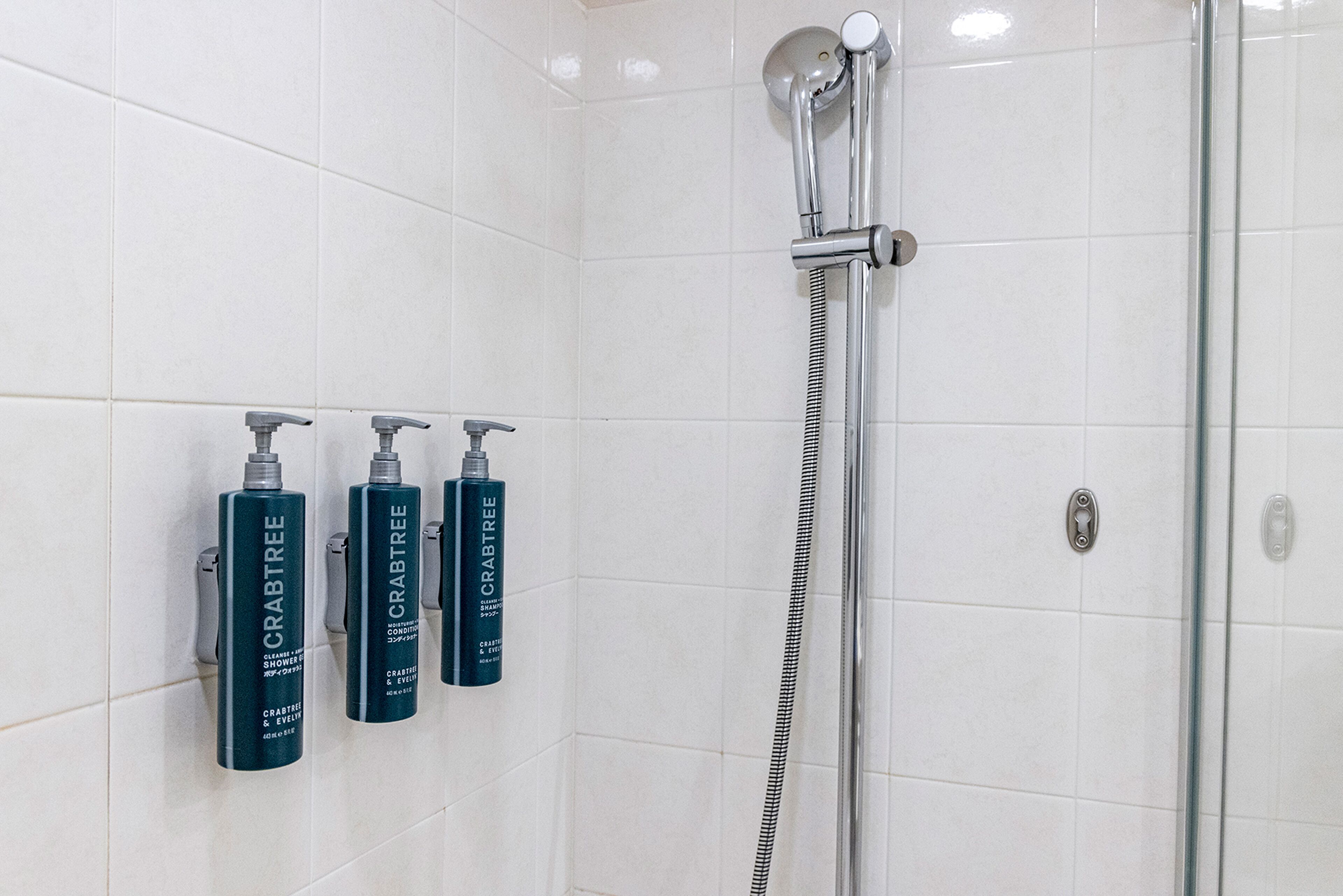 A shower area with a glass door, a handheld showerhead, and three bottles of Crabtree shower gel.