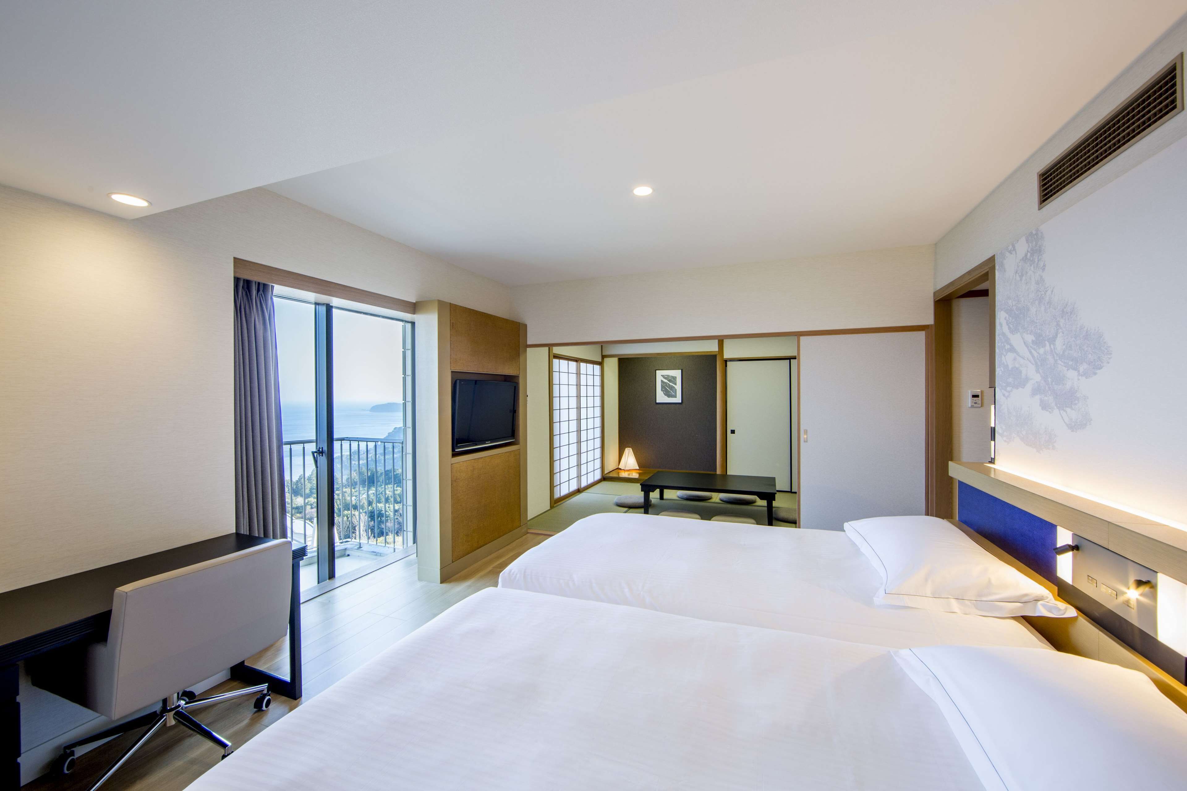 A hotel room with two beds, a desk, a TV, and a balcony with a view.