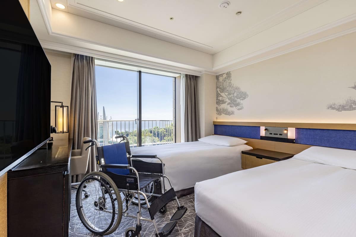 A hotel room with two beds, a wheelchair, a desk, and a TV.