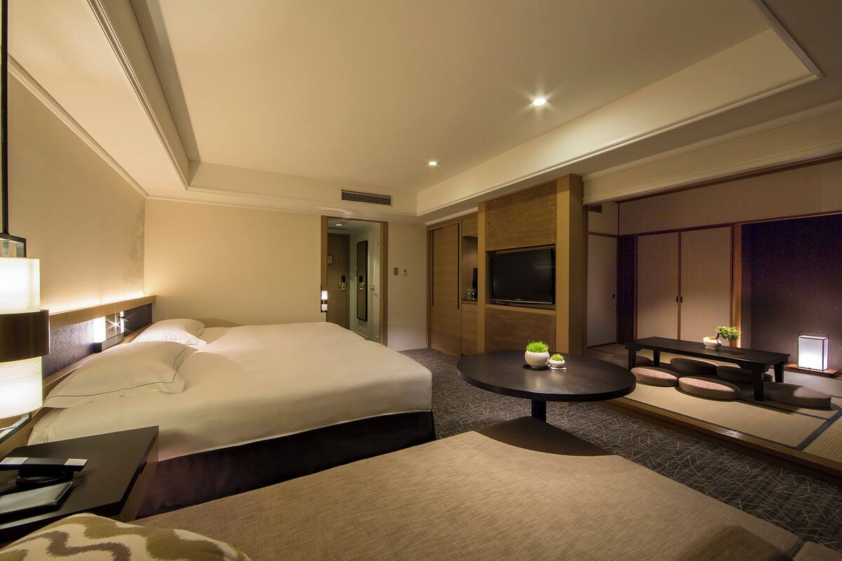A hotel room with a bed, bedside table, lamp, TV, and a small table with a plant.