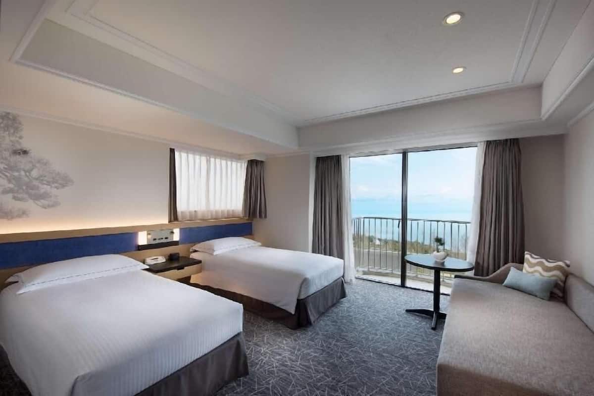 A hotel room with two beds, a small table, a sofa, and a balcony with a view.