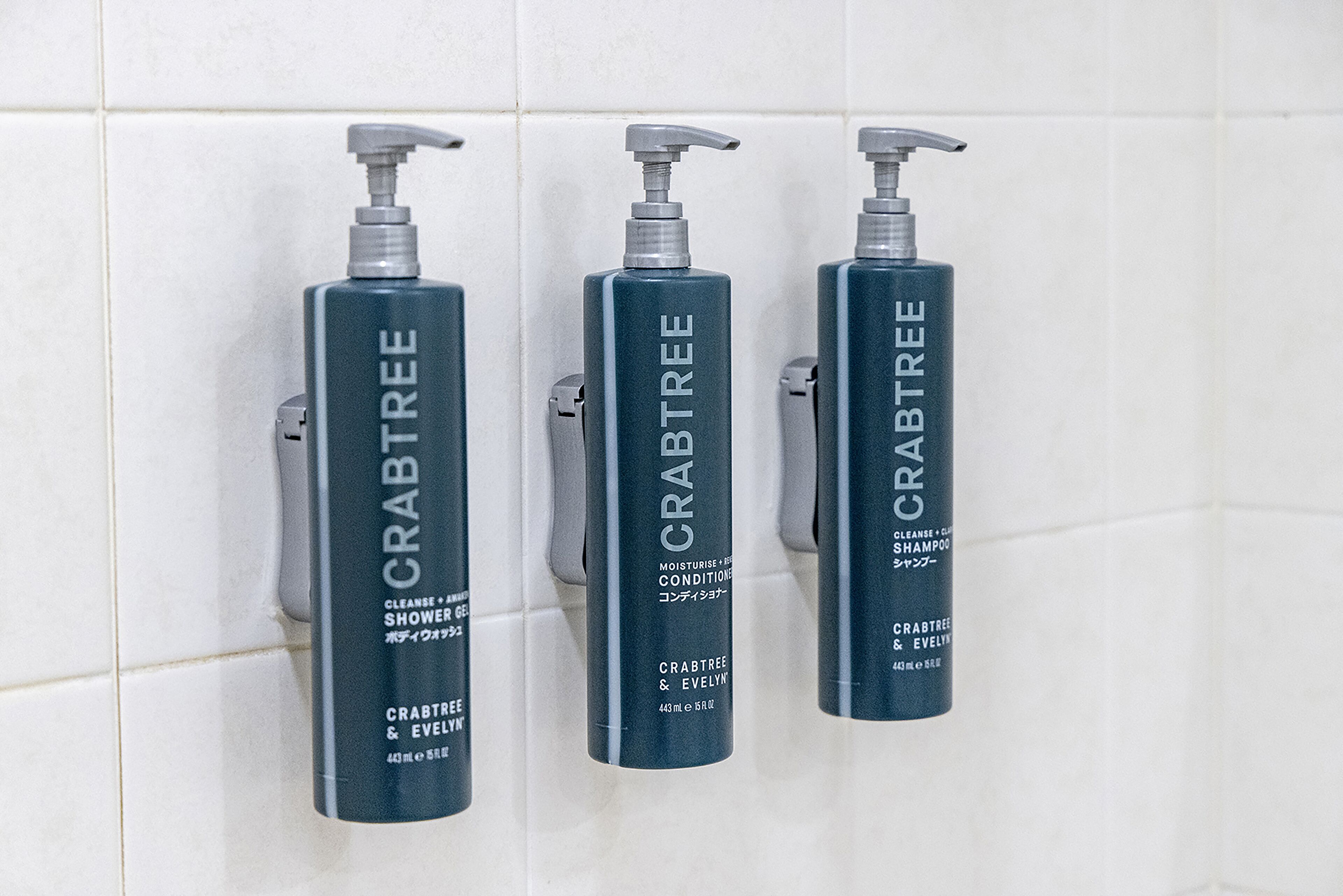 Three bottles of Crabtree & Evelyn shampoo and conditioner on a tiled wall.