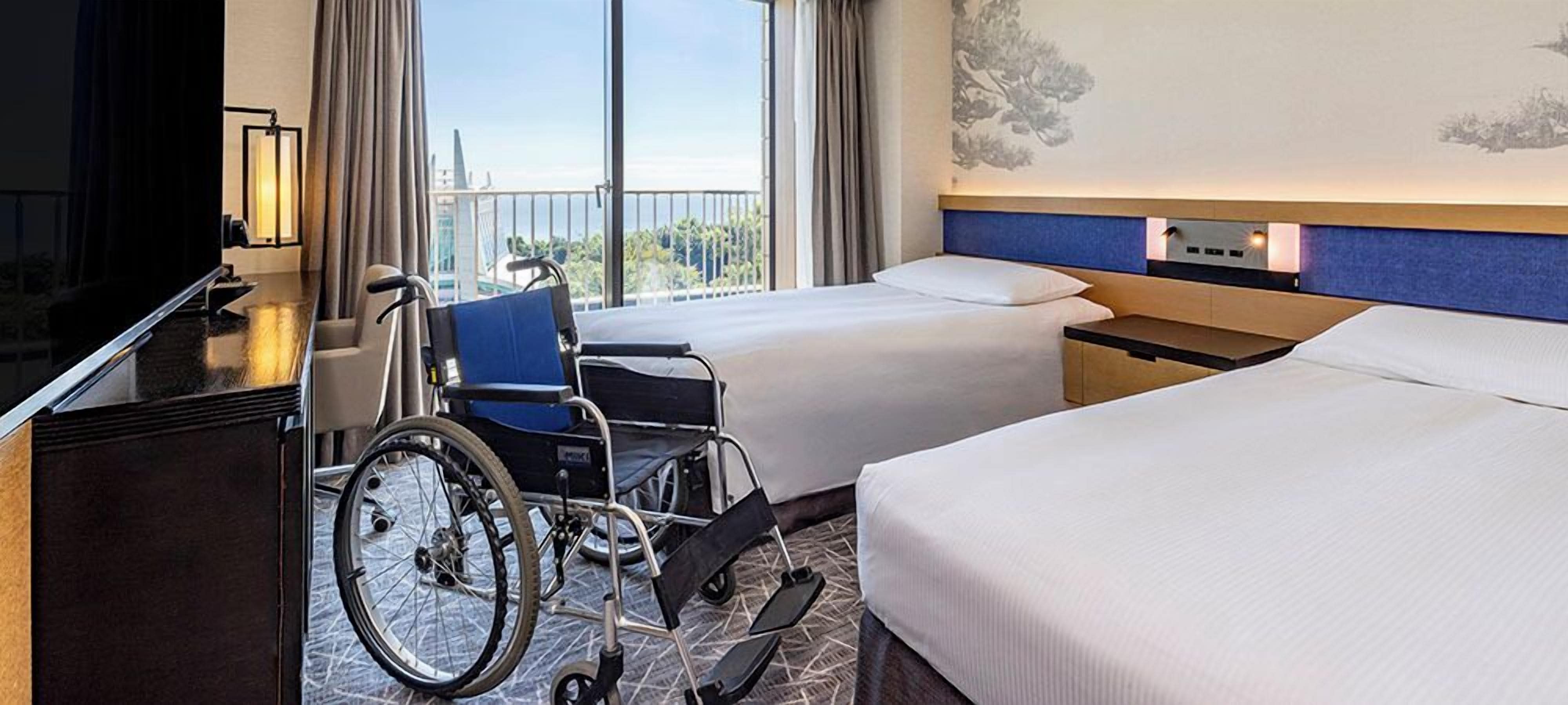A hotel room with two beds, a wheelchair, a desk, and a large window with a city view.