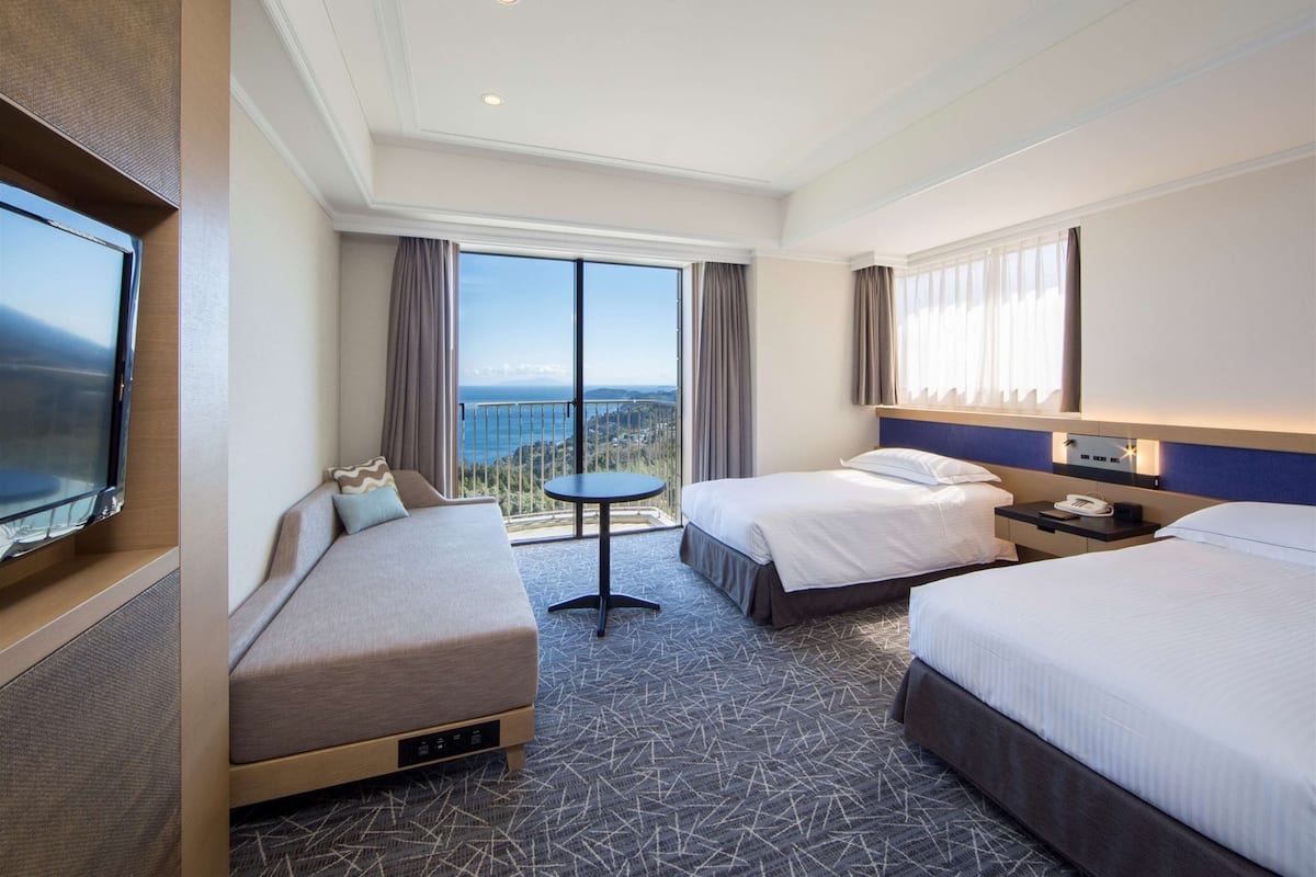 A hotel room with two beds, a TV, a small table, and a balcony with a view.