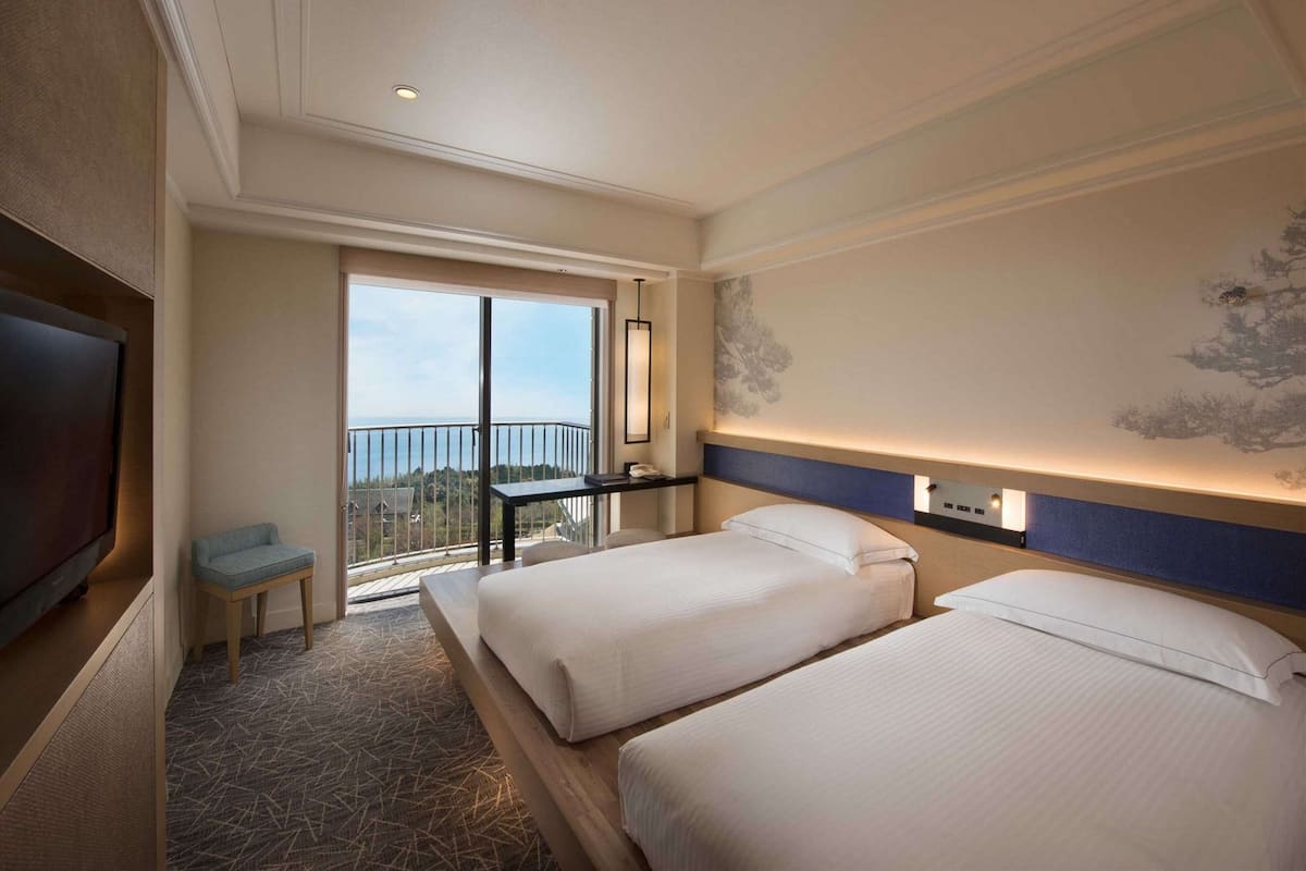 A hotel room with two beds, a TV, a balcony with a view, and a desk.