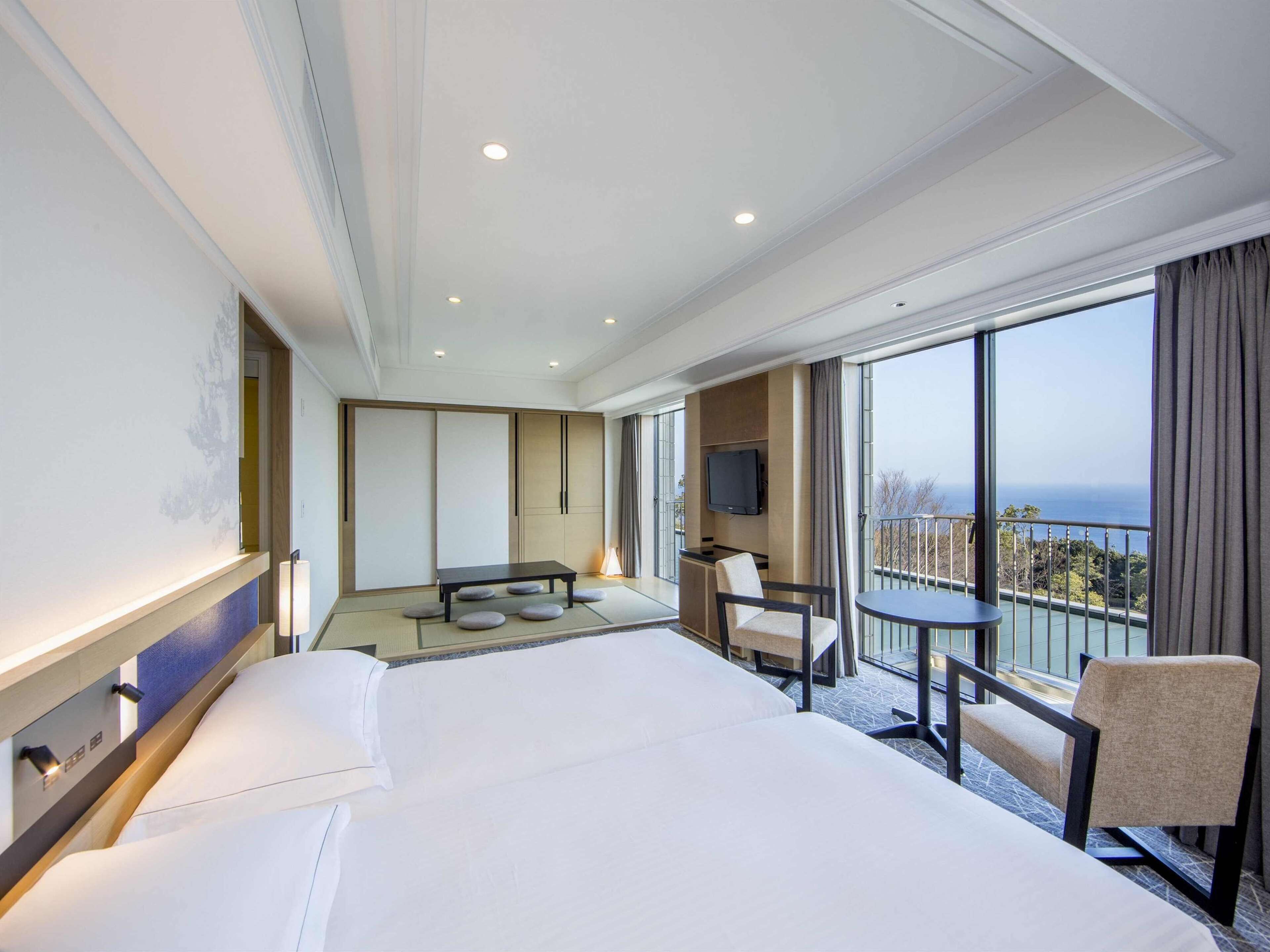 A hotel room with a large bed, a desk with a chair, a TV, and a balcony with a view.