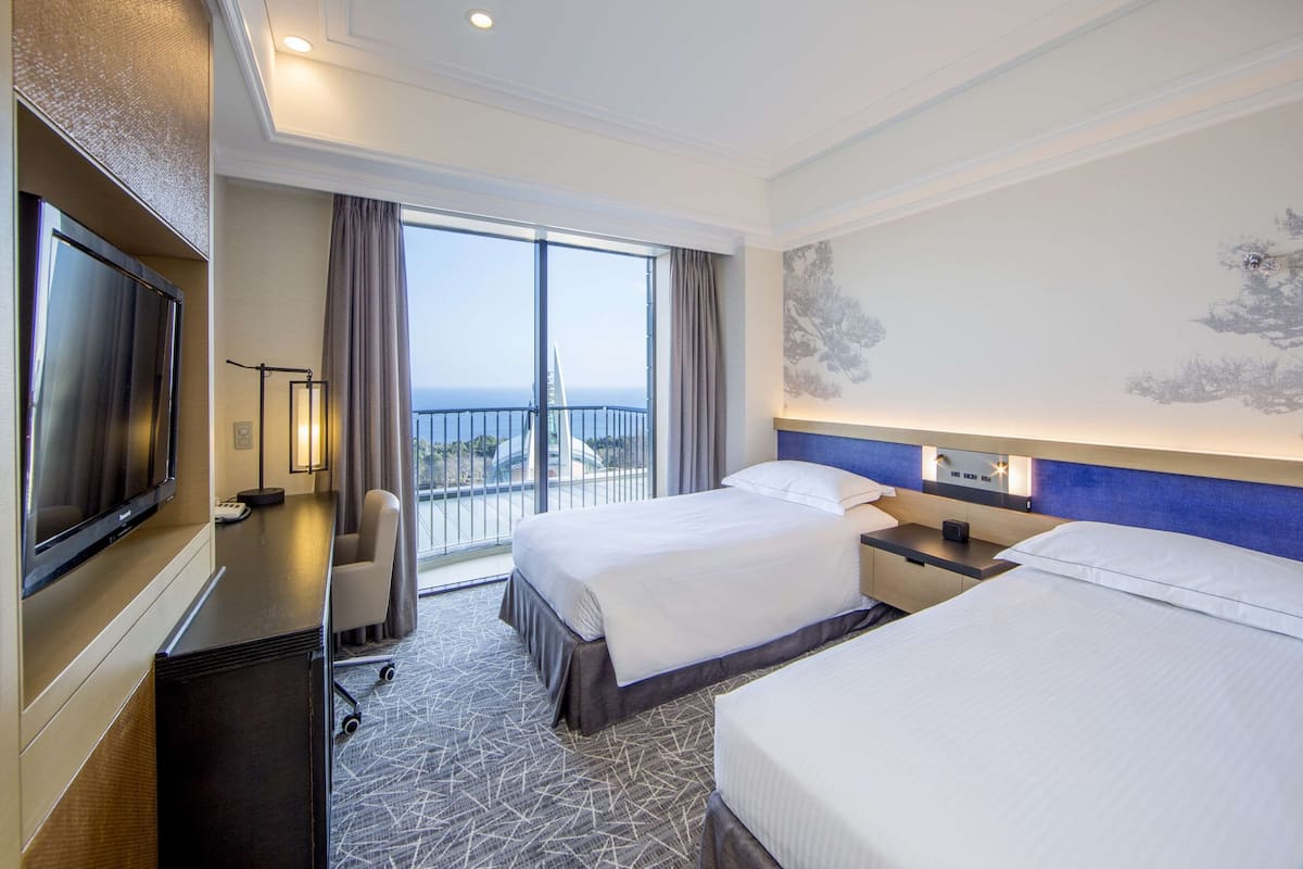 A hotel room with two beds, a desk, a TV, and a balcony with a view of the sea.