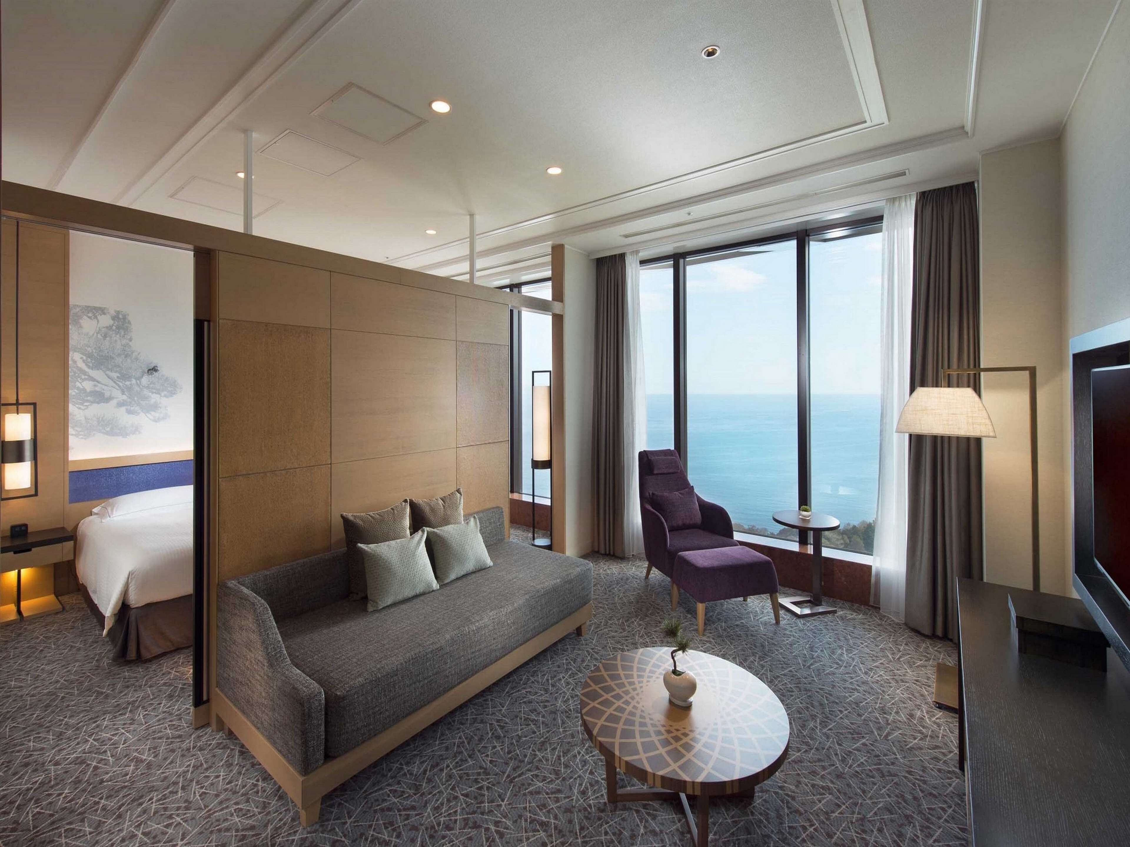 A modern hotel room with a large bed, a sofa, a chair, a small table, and a TV. The room has large windows with a view of the sea.