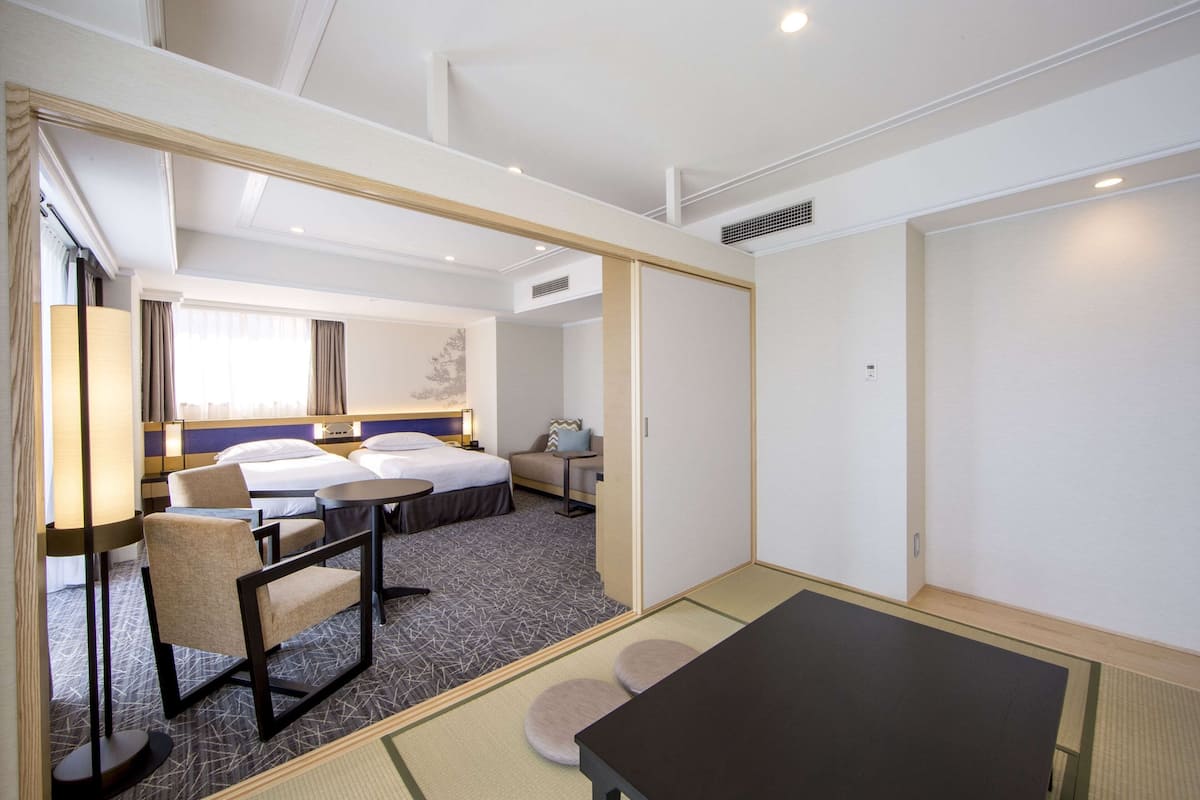 A hotel room with a bed, two bedside tables, a chair, a small table, and a view of the hallway.