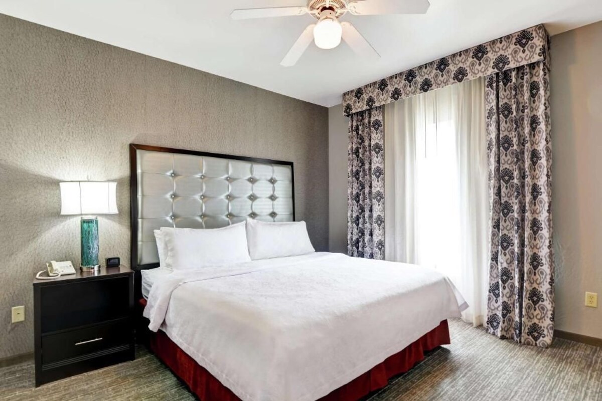 Une chambre d’hôtel avec un lit, une table de chevet, une lampe, un ventilateur de plafond et des rideaux à motifs.