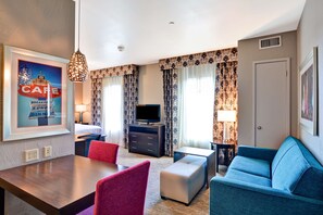 Habitación de hotel con una mesa de comedor, sillas rojas y azules, un sofá azul, un televisor y un cuadro en la pared.
