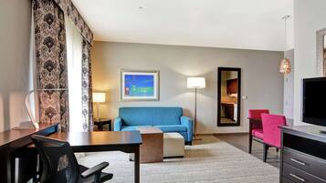 A hotel room with a desk, a blue sofa, a red chair, a TV, a lamp, and a framed picture on the wall.