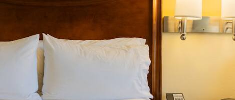 A neatly made bed with white linens and pillows, a wooden headboard, a bedside table with a lamp and a telephone, and a wall-mounted light fixture.