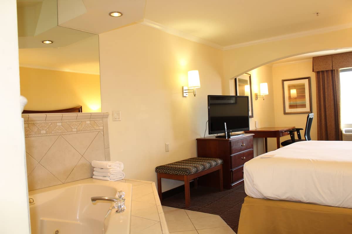 A hotel room with a large bed, a desk with a chair, a television, a bathroom with a bathtub, and a mirror.