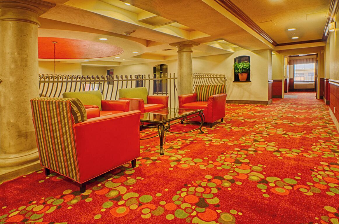 Un vestíbulo de hotel con alfombra roja, una zona de estar con sillas a rayas y un techo con columnas.