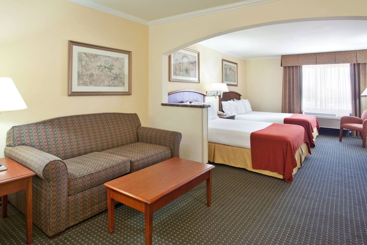 A hotel room with a sofa, a bed with red bedding, a desk, and a chair. There are framed pictures on the walls and a window with curtains.