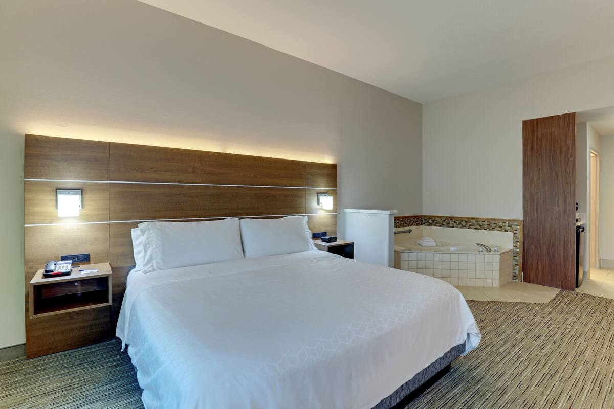 Habitación de hotel con una cama grande, una mesita de noche con lámpara, una mesita auxiliar y una bañera empotrada.