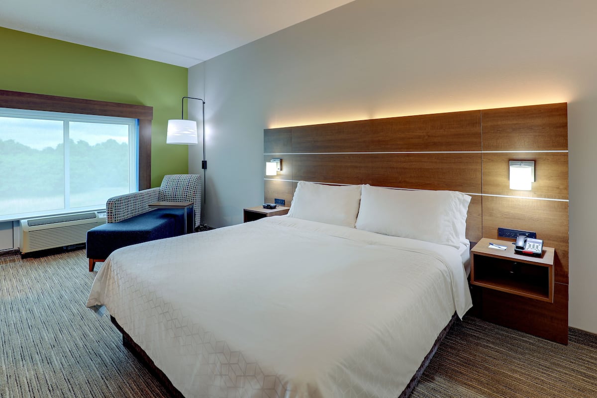 Habitación de hotel con una cama grande, una mesita de noche con lámpara, una silla y vistas al exterior.