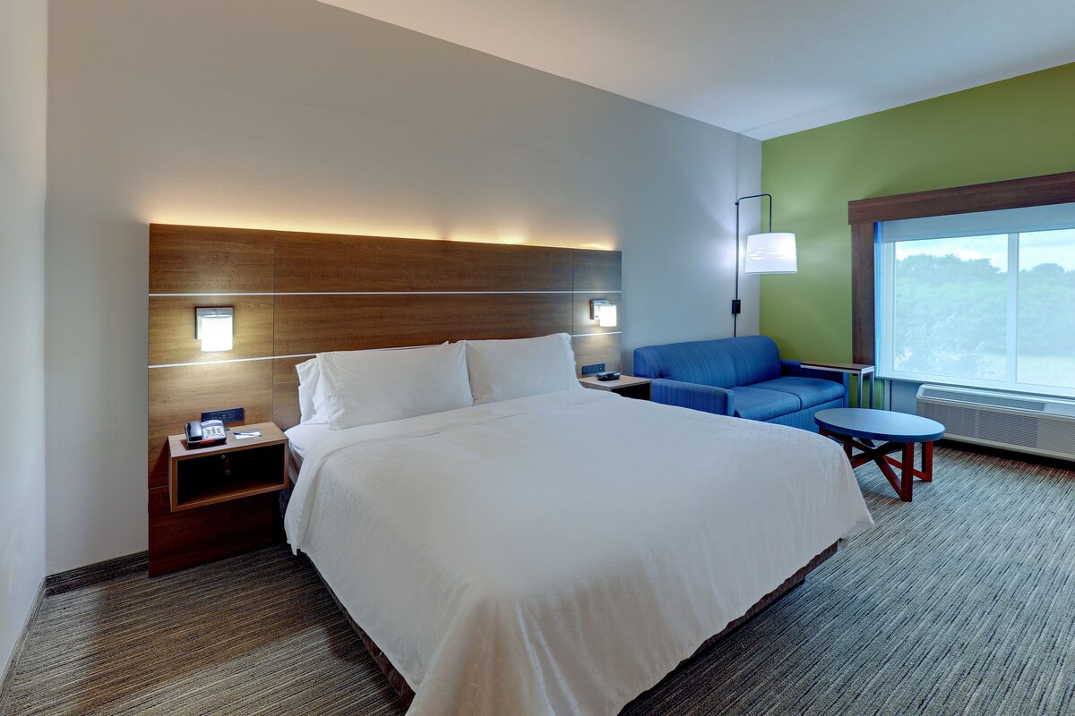 Habitación de hotel con una cama grande, una mesita de noche, un sillón azul, una pequeña mesa redonda y una ventana con vista a árboles.