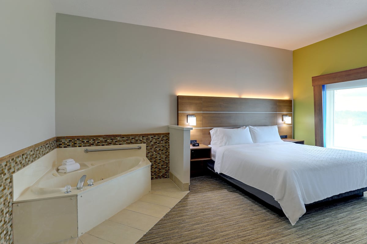 Habitación de hotel con una bañera grande, una cama con ropa blanca y una mesita de noche con lámpara.