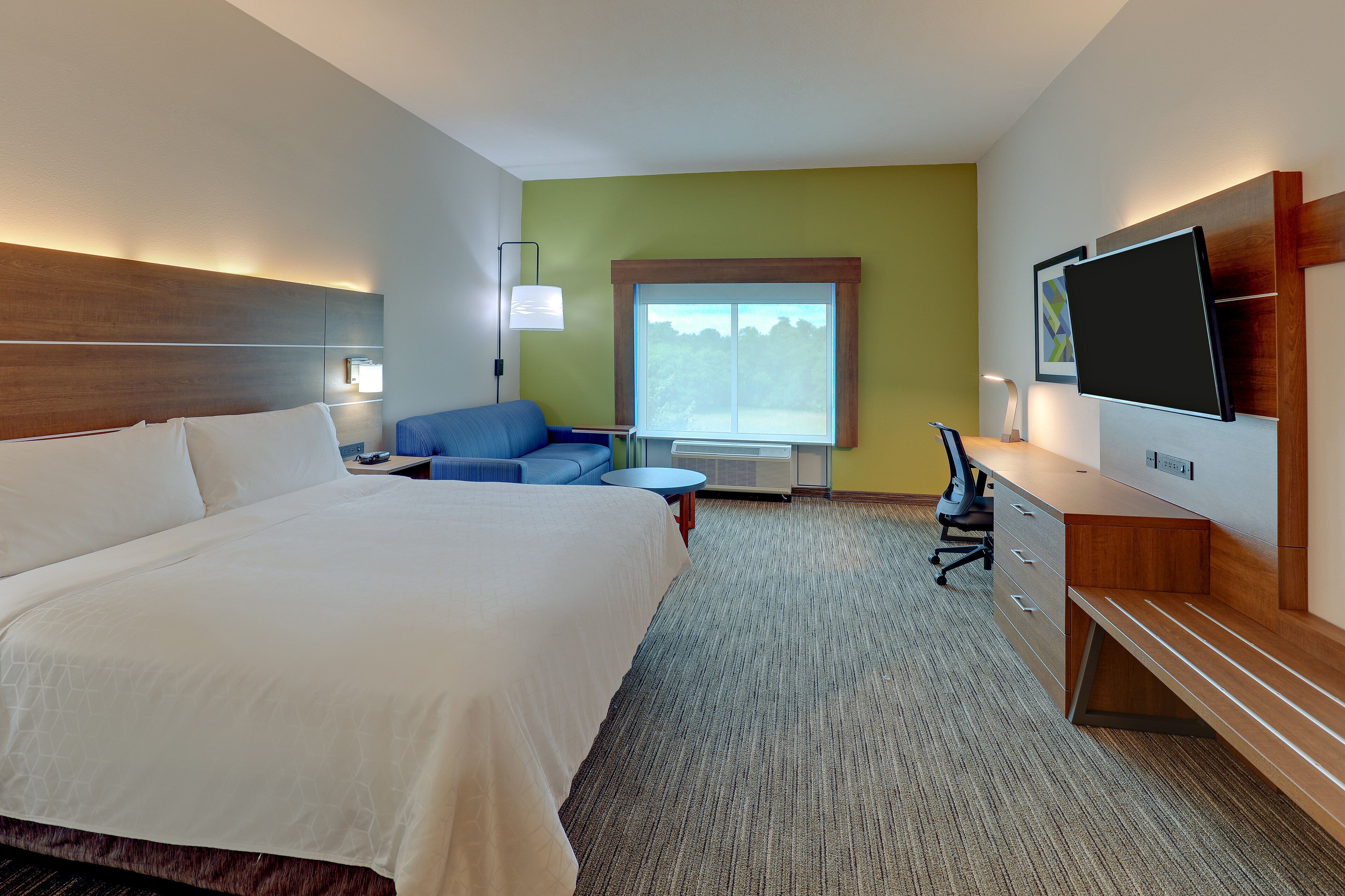 A hotel room with a large bed, a desk with a chair, a TV, and a window with a view of trees.