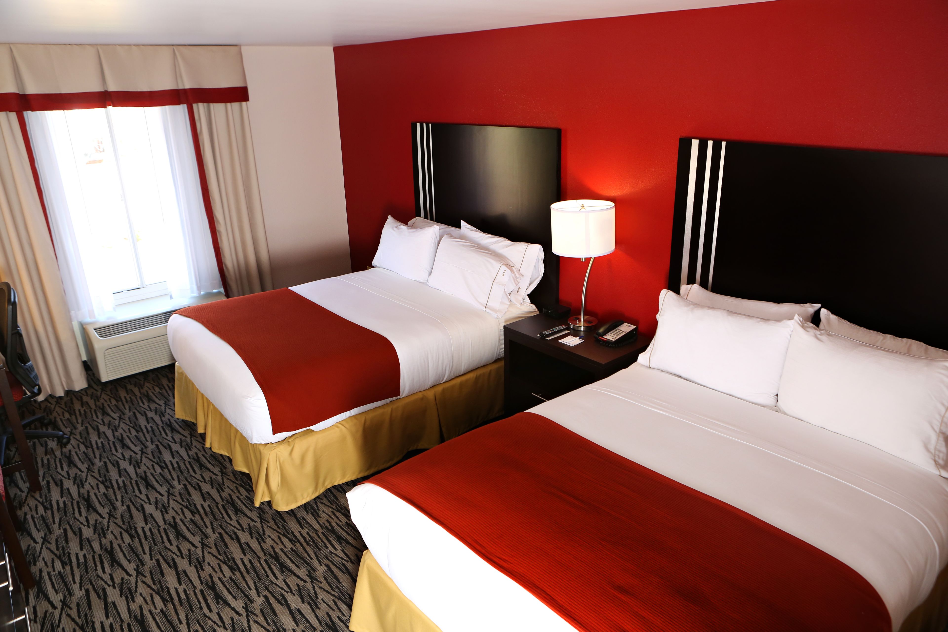 A hotel room with two beds, a red and white color scheme, a large window with curtains, and a carpeted floor.