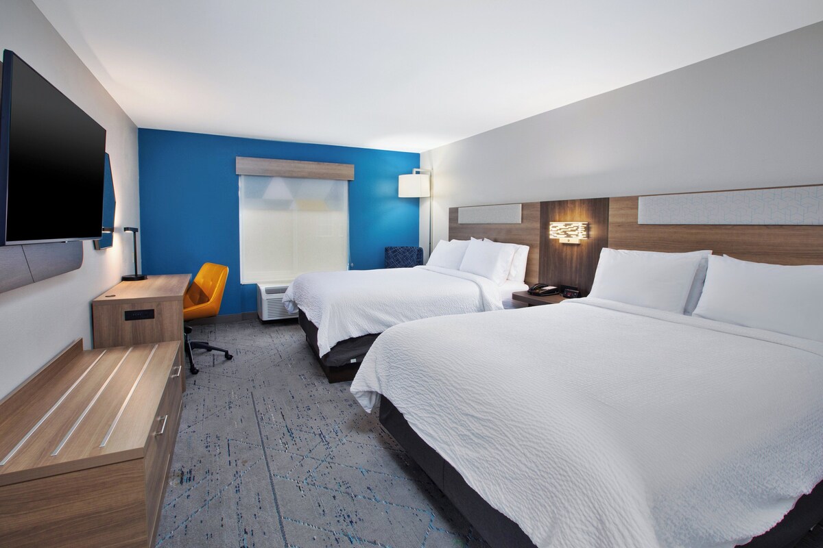 A hotel room with two beds, a desk, a chair, a flat-screen TV, and a window with blinds.