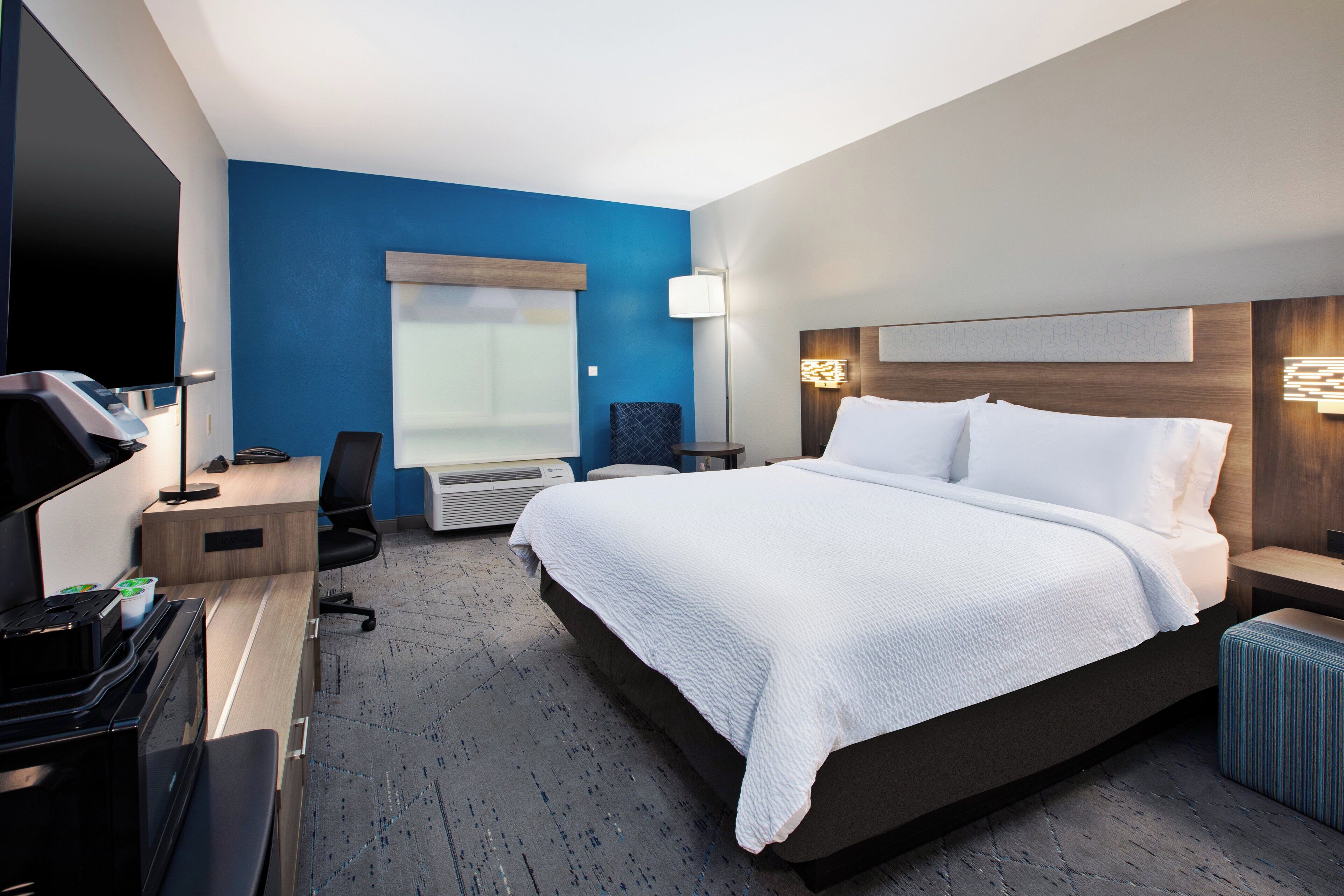 A hotel room with a large bed, a desk with a computer, a chair, a flat-screen TV, and a window with blinds.