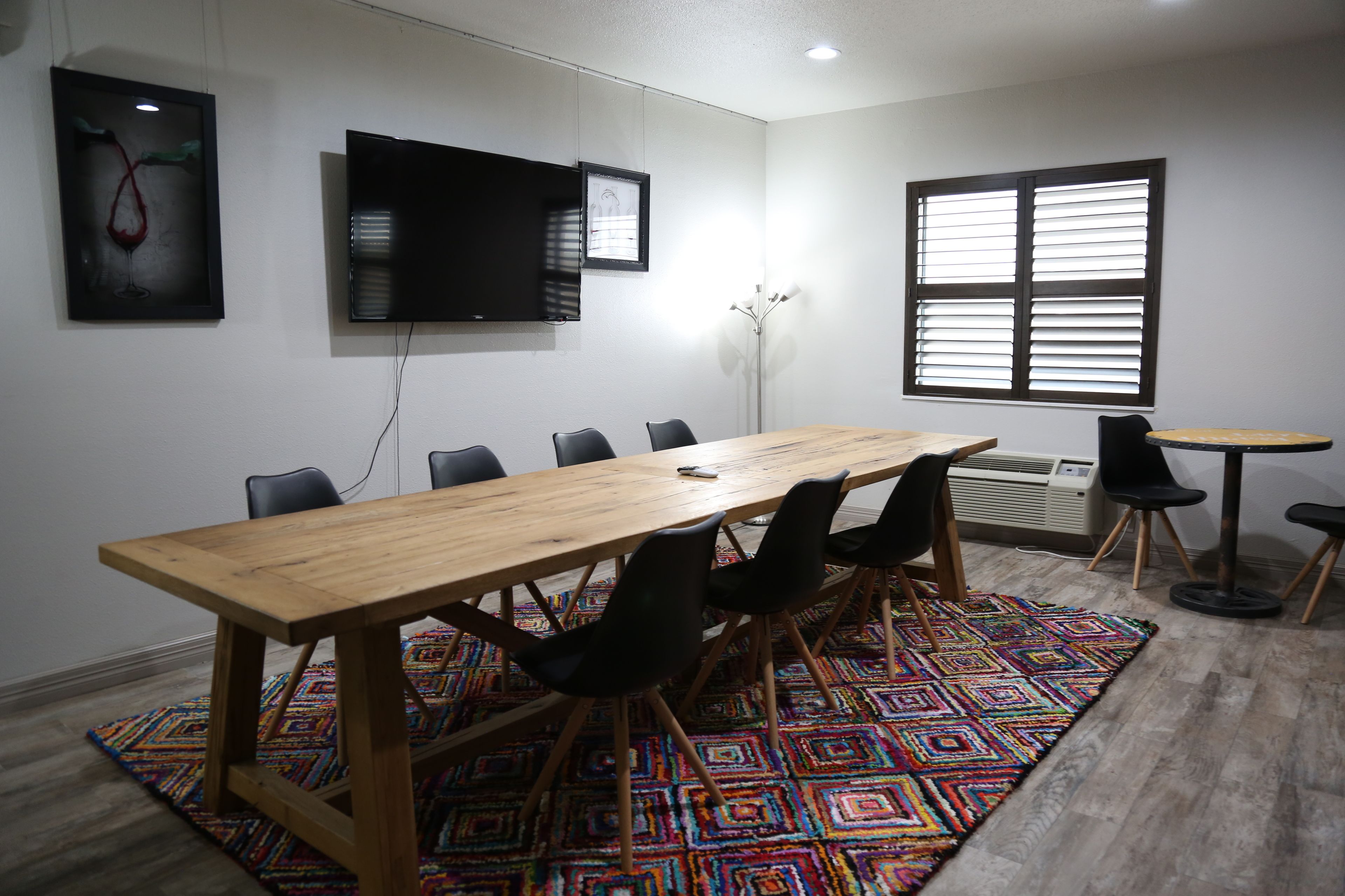 Uma sala de conferências com uma longa mesa de madeira, cadeiras pretas, um tapete colorido, uma TV fixada na parede e uma janela com persianas.