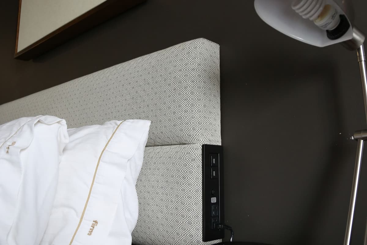 A hotel room with a bed, a bedside table with a lamp, a clock, and a framed quote on the wall.
