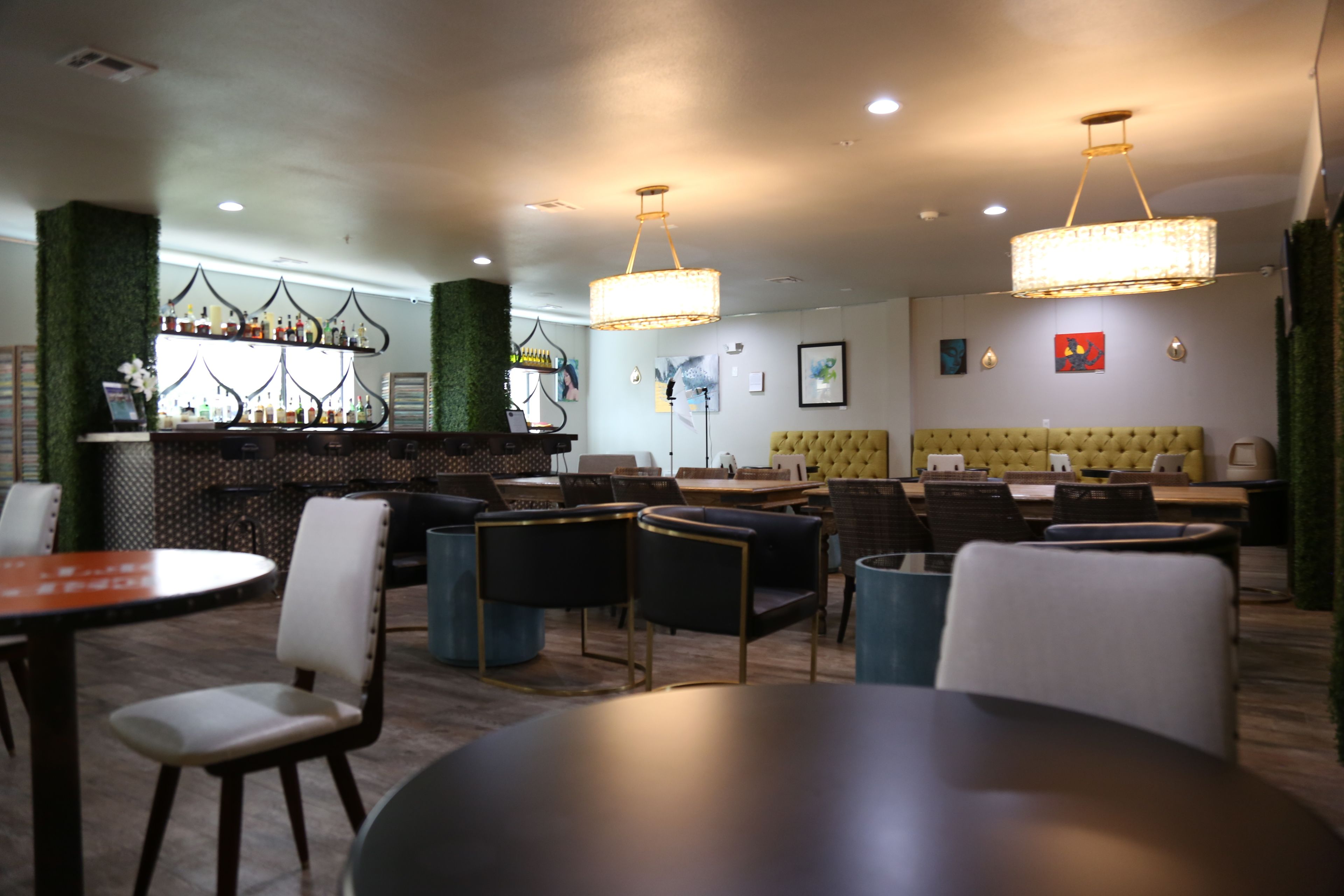 Un área de bar moderna con una pared verde, taburetes de bar y una variedad de botellas. Hay mesas redondas y sillas dispuestas para comer.