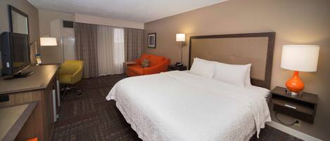 Ein Hotelzimmer mit einem großen Bett, einem Schreibtisch mit Computer, einem orangefarbenen Sessel und einem Fernseher.
