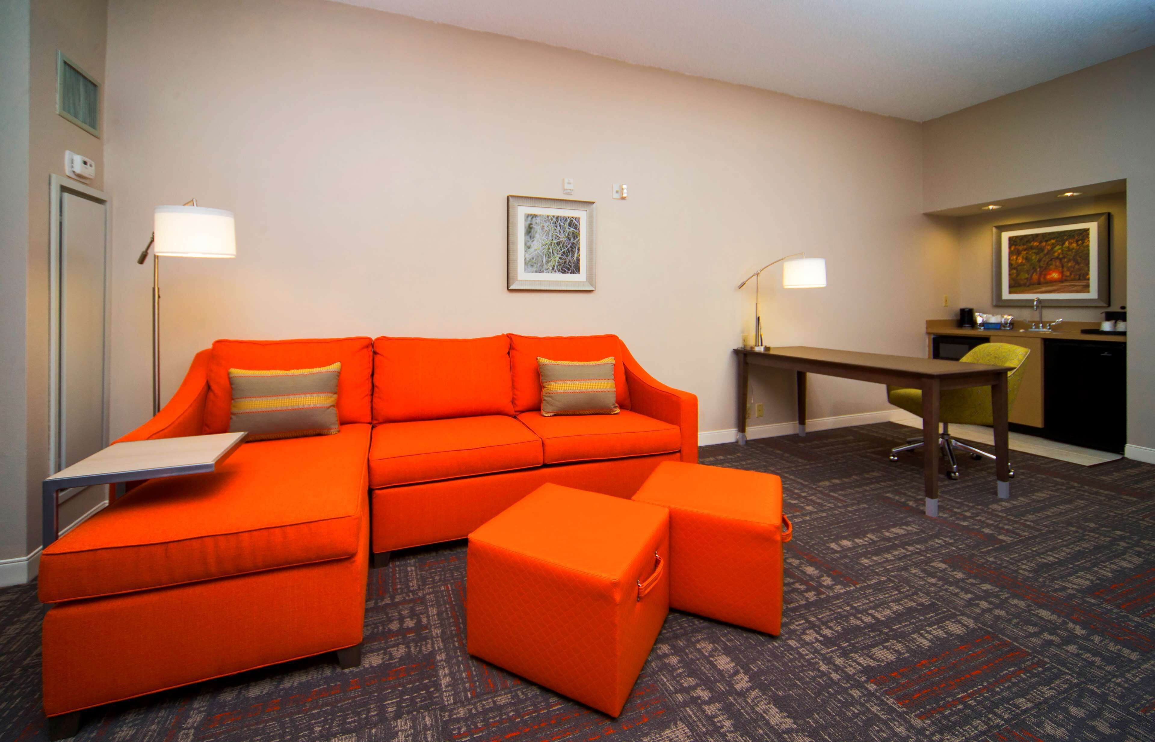 A modern hotel room with a bright orange sectional sofa, matching ottomans, a small desk, and a chair.
