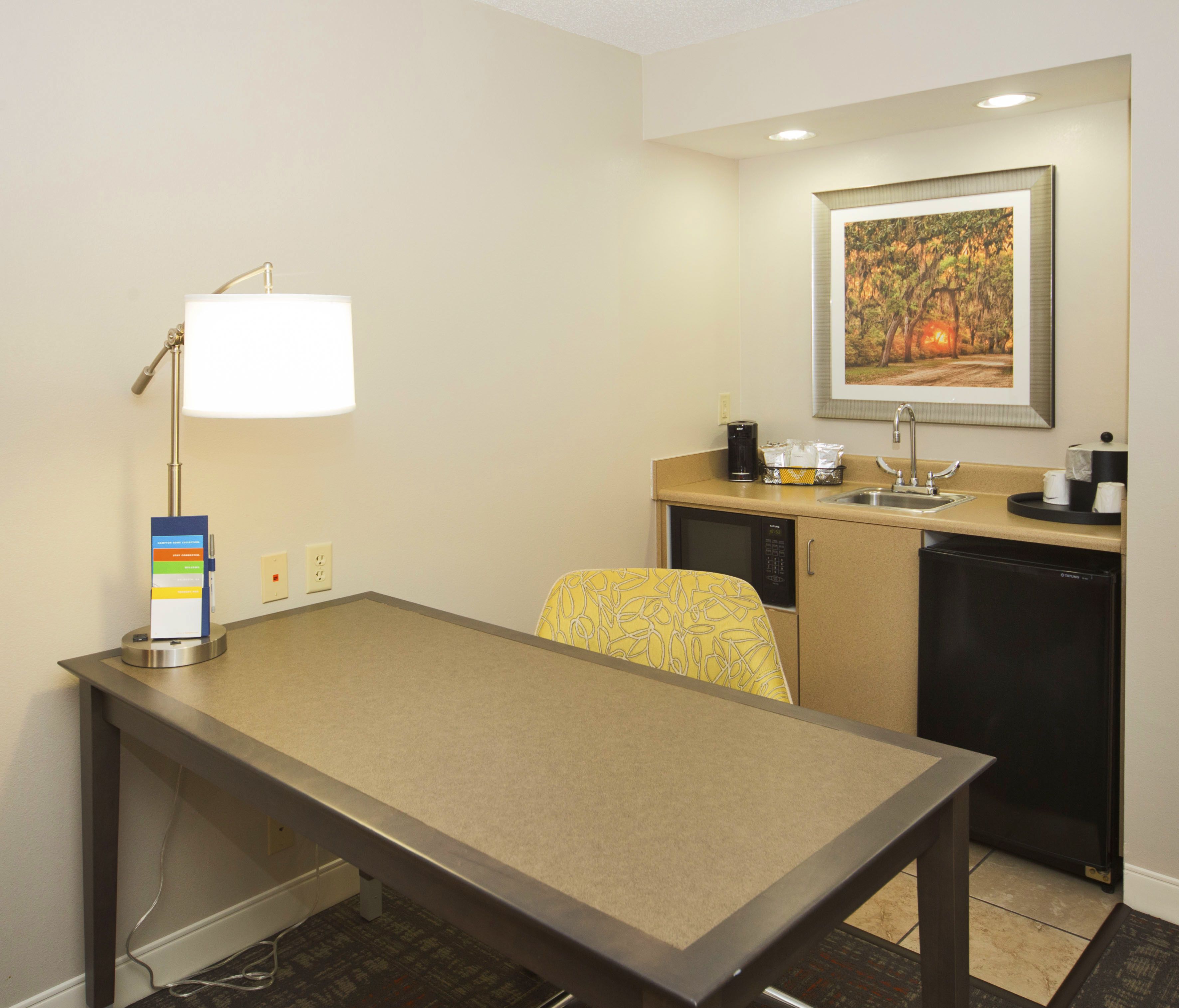 A hotel room with a kitchenette, a dining table, a chair, a lamp, a painting on the wall, and a small sink.
