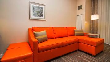 Quarto de hotel moderno com um sofá seccional laranja grande, uma mesinha e uma cama de casal.