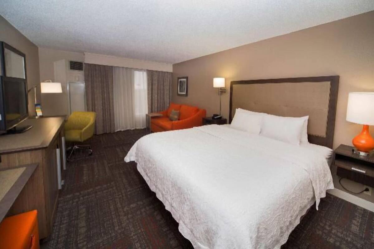 A hotel room with a large bed, a desk with a computer, an orange chair, and a TV.