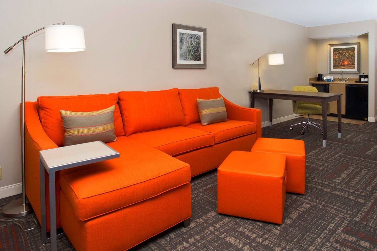 A modern hotel room with a bright orange sofa, matching ottoman, and a small table. There is a desk with a chair and a lamp in the background.