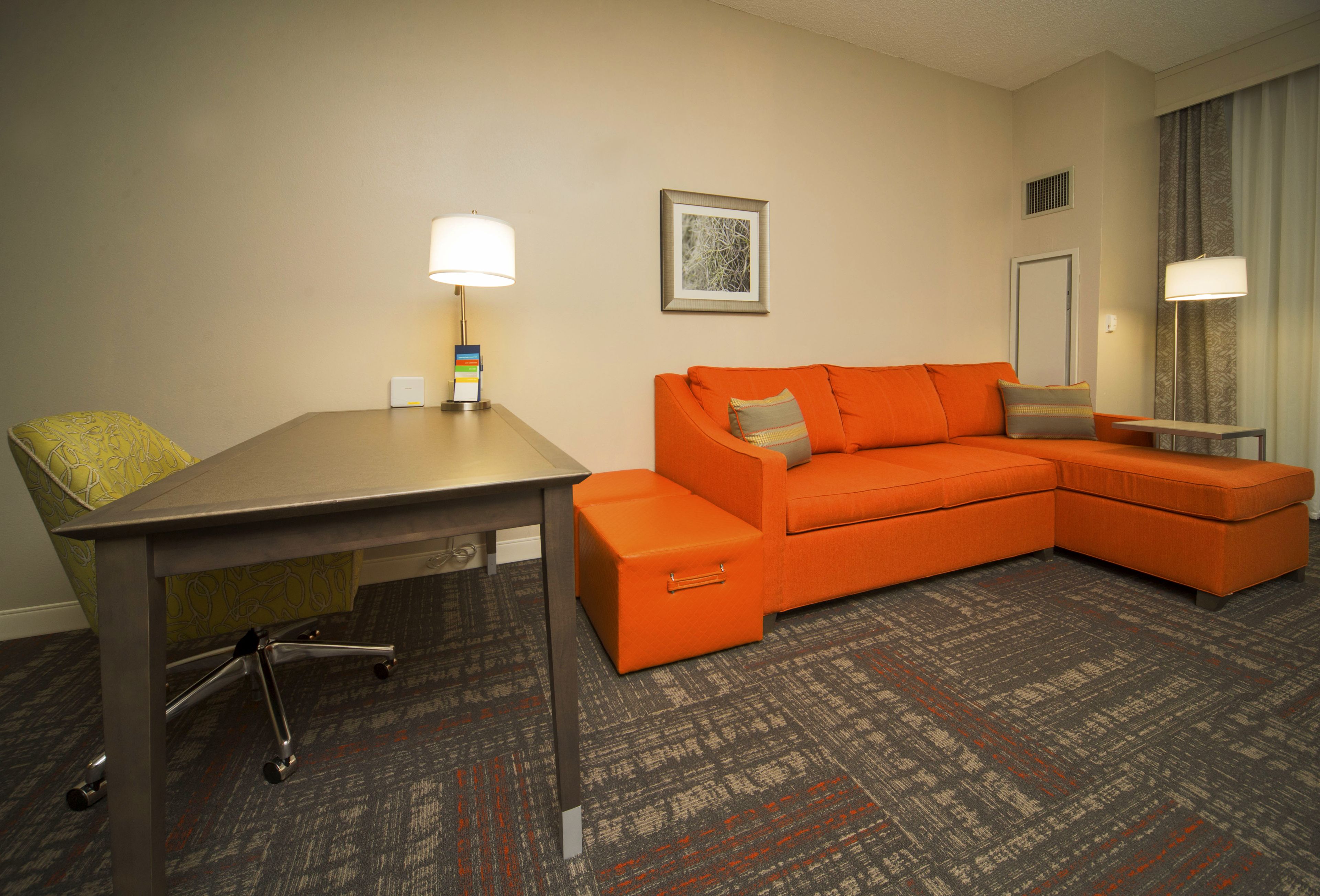 Une chambre d’hôtel avec un bureau, une chaise jaune, un canapé orange et un tapis à motifs.