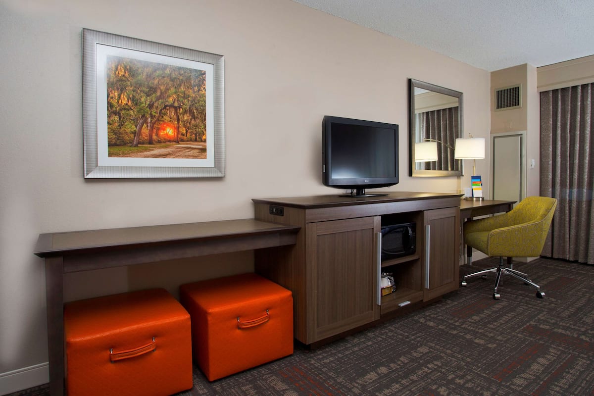 A hotel room with a desk, a television, a chair, and a framed picture on the wall.