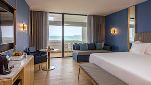 A hotel room with a large bed, a desk with a coffee machine, a small table with drinks, a sofa, and a view of the ocean.