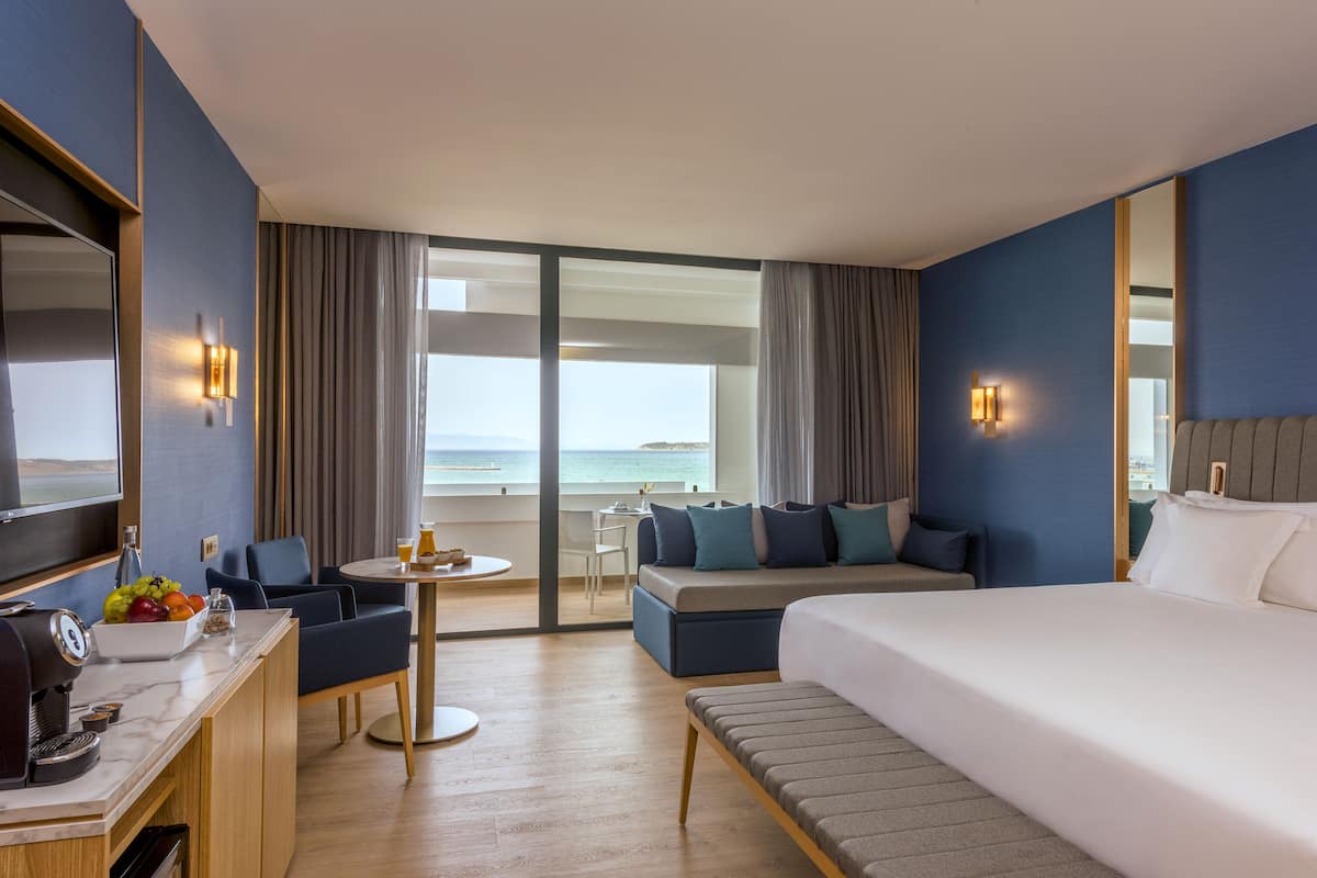 A hotel room with a large bed, a desk with a coffee machine, a small table with drinks, a sofa, and a view of the ocean.