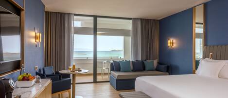 A hotel room with a large bed, a desk with a coffee machine, a small table with drinks, a sofa, and a view of the ocean.