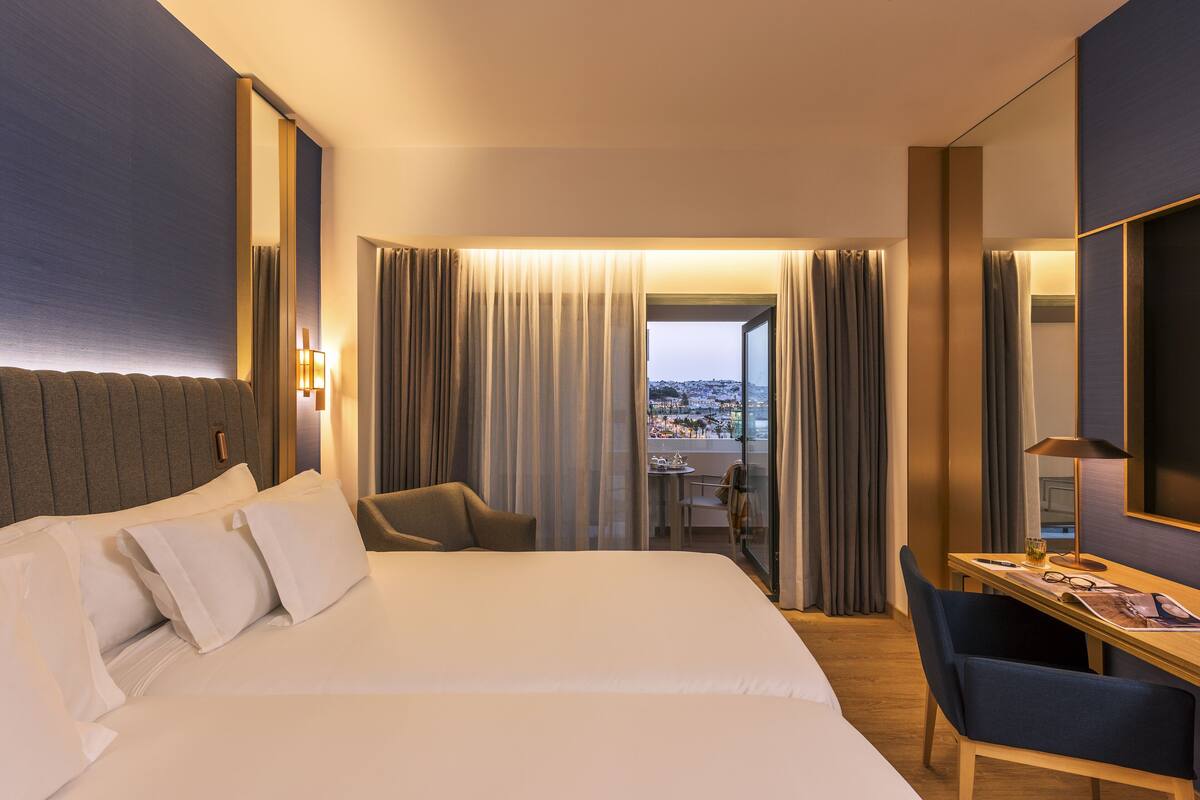 A hotel room with a large bed, a desk, a chair, and a view of a cityscape through the window.