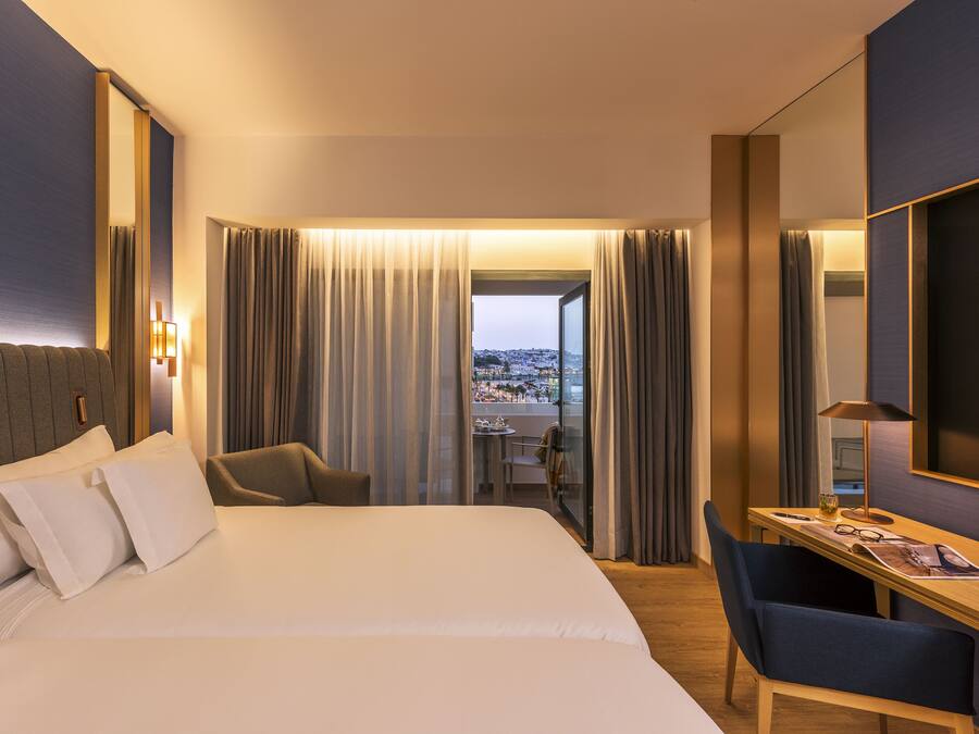 Habitación de hotel con una cama grande, un escritorio, una silla y vistas de la ciudad a través de la ventana.