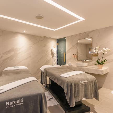 A modern spa room with two massage tables, a blue chair, a sink, and a mirror.