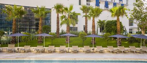 A swimming pool with lounge chairs and umbrellas, surrounded by palm trees and a modern building with balconies.
